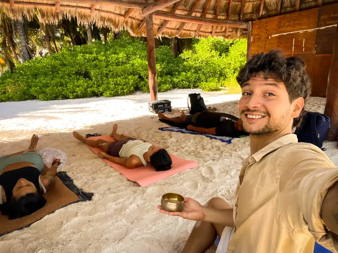 Tomi sonriendo con un cuenco en la mano mientras sus alumnas descansan en la arena